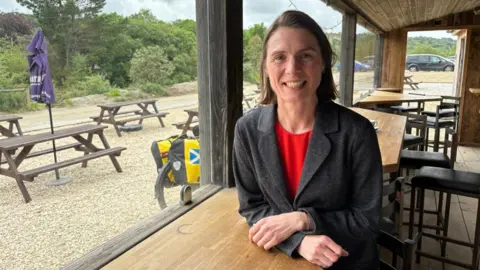 Lee Trewhela/LDRS Ruth Gripper sat at a table. She has short brown hair and is wearing a red top and grey coat. Behind her are tables and stool chairs. To the left of her is a window with a pub garden, it has benches and umbrellas.