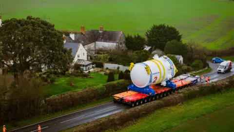 EDF A cylinder reactor which is as tall as a two-storey home next to it is being transported along a road in rural Somerset. The transporter takes up both lanes of the road.