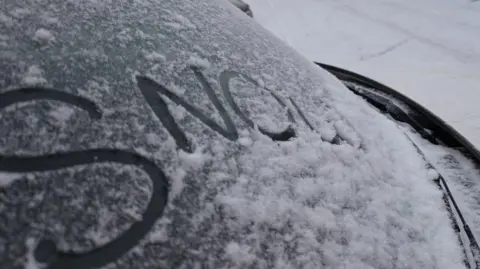 Lucie Johnson The rear window of a car covered in snow. Someone has written the word
