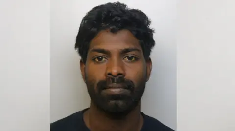 Avon and Somerset Police A police mugshot of Manoj Chinthathira wearing a black t-shirt and standing against a grey background. He has brown eyes, a full black beard and thick wavy black hair.