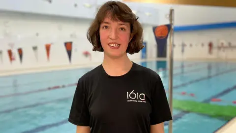 BBC Emily Gold. She has dark short hair and is pictured standing in front of a leisure centre swimming pool. She is wearing a black t-shirt with 1610 on the chest. She is looking directly at the camera and smiling.