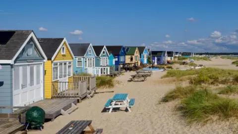 Getty Images A row of brightly coloured beach huts stand on a sandy beach next to some small dunes. There are picnic benches outside the huts. The sky is blue and it is a sunny day.