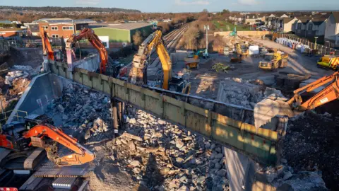 North Somerset Council Diggers and other construction machinery are surrounded by rubble as a bridge is demolished over a railway track. It is a sunny winters day in a residential area.