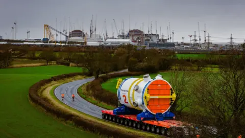 EDF A huge white, orange and yellow cylinder reactor is being transported on a wide lorry base in the foreground. Two people in orange high-visibility clothes guide it along the route. There's a narrow road leading up to the Hinkley Point C construction site in the background, which is filled with dozens of cranes and has two central domes.