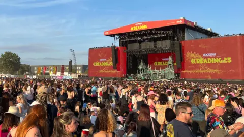 BBC An enormous crowd of people stand in front of a large stage. Massive red signs either side of the stage read