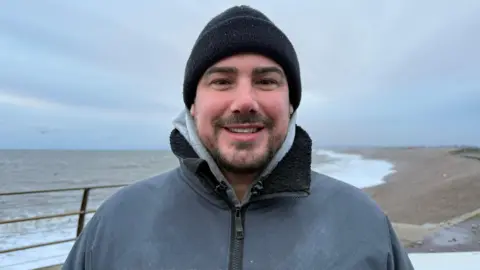 BBC A man smiling wearing a black hat and grey rain coat on the beach.