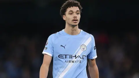 Getty Images Max Alleyne during a Premier League match. He has curly brown hair and a dark brown moustache. He is wearing a Manchester City kit which is light blue and has a white diagonal stripe from one hip to one shoulder. The background is blurred and is very dark. He is lit up by bright lights.
