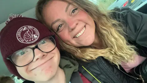 Family photo Harry Basham and his mum Leanne Golding are smiling at the camera. They are sitting inside a car. Harry is wearing a burgundy hat with Taunton Town Football Club written on it. Leanne is wearing a jacket and a pink top. She has long wavy hair.